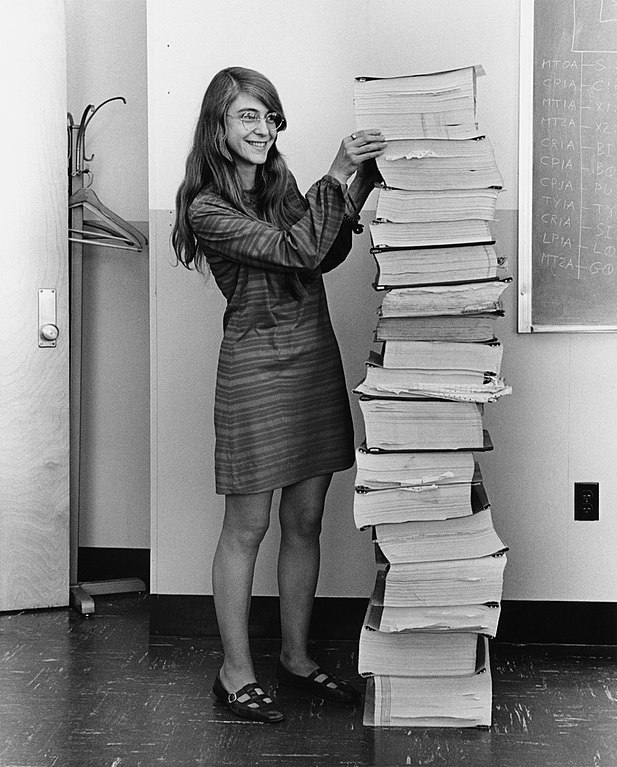 Margaret Hamilton đứng bên chồng giấy ghi lại đoạn mã của phần mềm điều hướng mà nhóm của bà tại MIT đã phát triển cho chương trình Apollo. Ảnh: Wikimedia Commons.